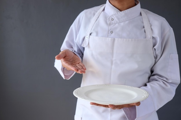 Image recadrée d'un chef masculin en tablier blanc tenant une assiette vide avec le geste de présentation du bras.