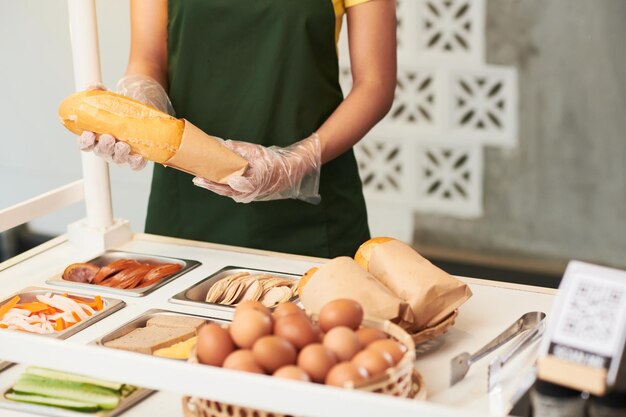 Image recadrée d'un cuisinier dans des gants jetables préparant un sandwich et l'emballant dans un emballage en carton