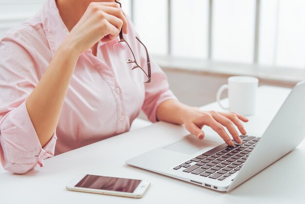 Image recadrée d'une femme d'affaires en chemise classique utilisant un ordinateur portable et tenant des lunettes tout en travaillant au bureau