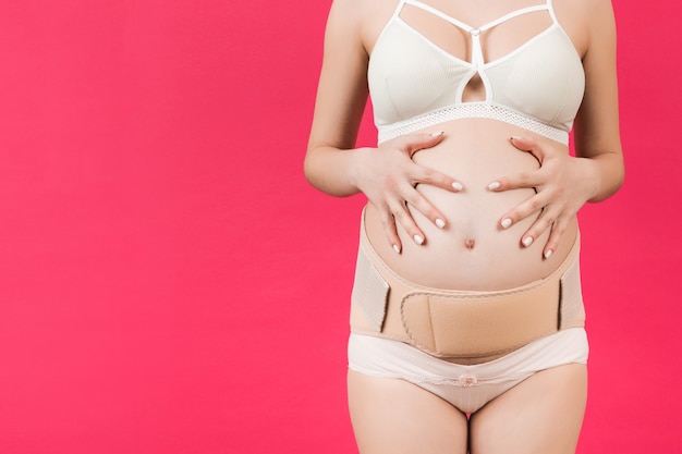 Image recadrée d'une femme enceinte en sous-vêtements portant un bandage élastique contre la douleur dans le dos sur fond rose avec espace de copie. Concept de ceinture de soutien abdominale orthopédique.