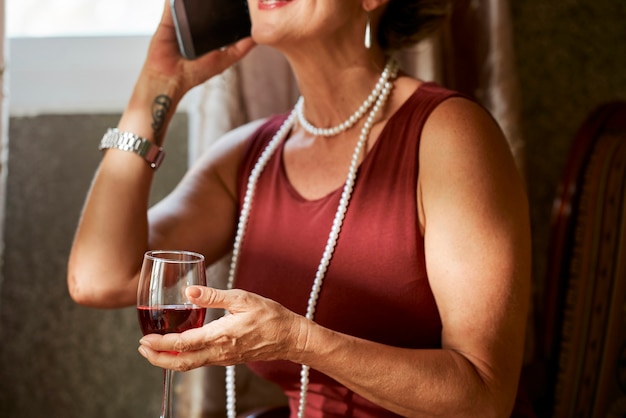 Image recadrée d'une femme mûre avec un collier de perles buvant du vin rouge et parlant au téléphone