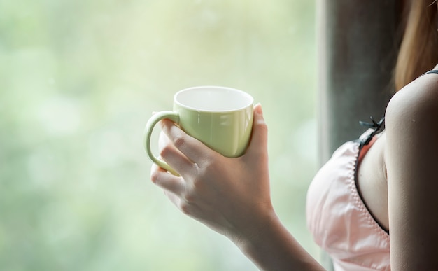 Image recadrée de femme sexy tenant une tasse de café debout près de la fenêtre