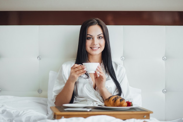 Image recadrée d'une fille brune heureuse assise dans son lit avec un bouquet de roses, mangeant un croissant avec du café sur un plateau le matin