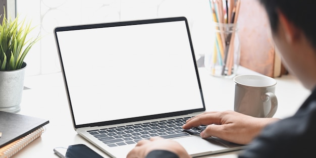 Image recadrée d'homme d'affaires en tapant sur un ordinateur portable à écran blanc blanc qui met sur le bureau avec une tasse de café, plante en pot, porte-crayon, livres. Concentré de concept de travail.