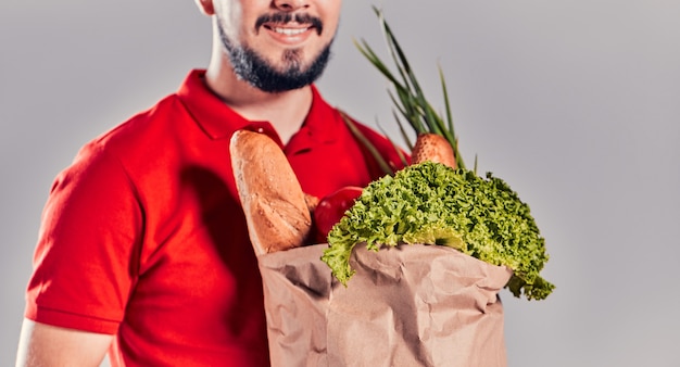 Image recadrée d'un jeune coursier barbu vêtu d'un T-shirt rouge et d'une casquette avec un paquet de nourriture. Nourriture livrée à domicile.