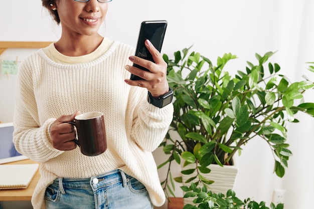 Image recadrée d'une jeune femme entrepreneur debout dans un bureau à domicile, buvant une tasse de café et lisant des messages texte sur un smartphone