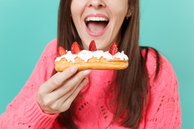 Image recadrée d'une jeune femme en pull rose tricoté tenir dans la main en train de manger, de mordre le gâteau éclair isolé sur fond de mur bleu turquoise, portrait en studio. Concept de mode de vie des gens. Maquette de l'espace de copie.