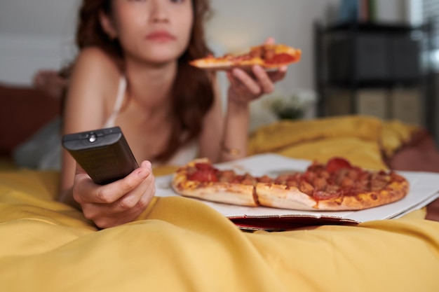 Photo image recadrée d'une jeune femme se relaxant sur son lit à la maison, mangeant de la pizza et regardant une émission à la télévision