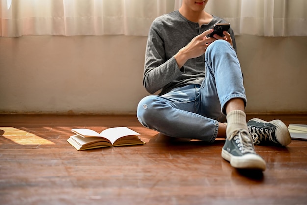 Image recadrée d'un jeune homme asiatique assis par terre dans sa chambre défilant sur son téléphone