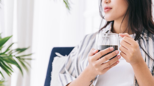 Image recadrée des mains d'une femme tenant une tasse de café dans le salon avec une plante verte Se détendre en buvant une tasse de café chaud et en profitant d'un moment calme et d'un environnement intérieur dans une maison confortable