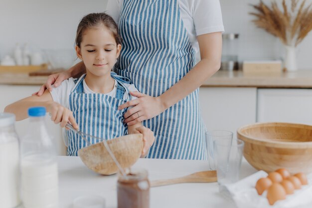 Image recadrée de mère affectueuse en tablier embrasse sa fille qui apprend à faire du pâteux