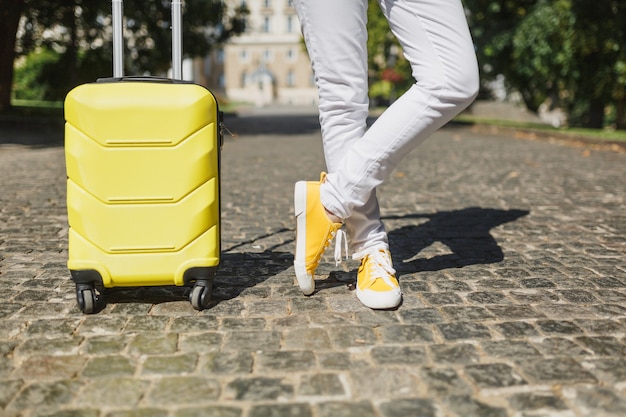 Image recadrée voyageur touriste femme jambes croisées dans des vêtements décontractés d'été jaune avec valise sur route en plein air de la ville. Fille voyageant à l'étranger pour voyager le week-end. Mode de vie de voyage touristique.