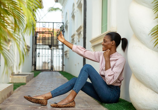 Image d'une seule fille africaine assise prenant un selfie en plein air avec enthousiasme