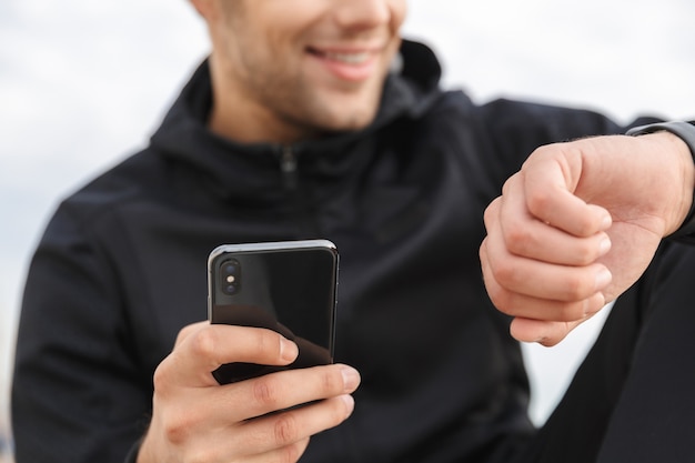 Image de sourire sportif de 30 ans en vêtements de sport noir, à l'aide de smartphone assis sur une promenade au bord de la mer