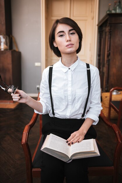 Image verticale de l'auteur en chemise blanche assise sur une chaise avec un livre sur les jambes