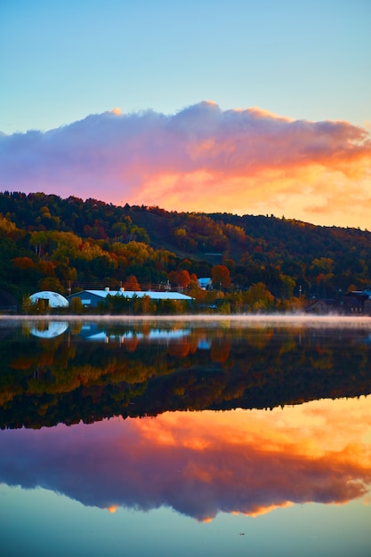 Image de la verticale d'un ciel bleu lever de soleil avec des nuages violets roses et oranges avec un miroir d'eau et une maison ou une grange avec des arbres d'automne