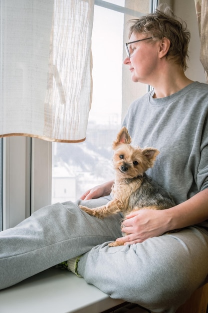Image verticale d'une femme d'âge moyen avec une coupe de cheveux courte et des lunettes avec un chien sur ses genoux Une femme et un petit chien sur le rebord de la fenêtre regardent par la fenêtre