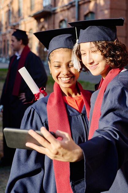 Image verticale de filles en robes de graduation faisant un selfie portrait sur un smartphone à l'extérieur