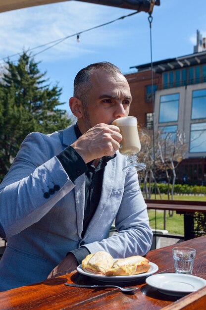 Image verticale d'un homme d'affaires adulte exécutif vêtu de vêtements élégants, assis le matin, prenant son petit-déjeuner et buvant du café à l'extérieur de l'espace de copie du restaurant