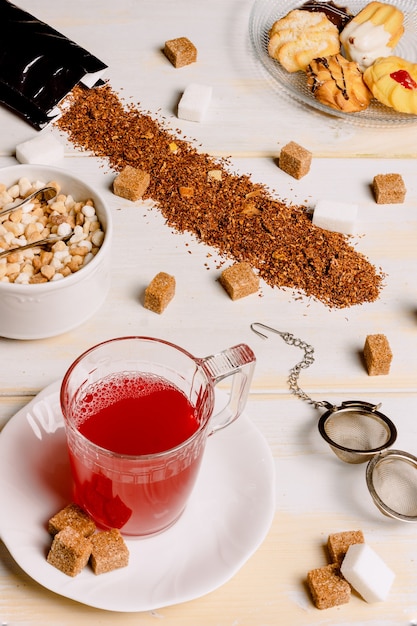 Image verticale d'une tasse de thé rouge sur des planches de bois blanc patiné avec des cubes de sucre éparpillés sur la table et la plaque avec des pâtisseries à thé dans le coin supérieur