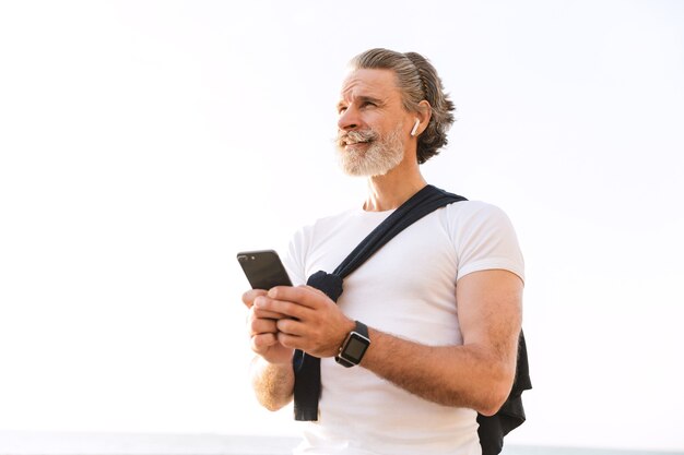 Image d'un vieil homme heureux en tenue de sport utilisant des écouteurs et un téléphone portable tout en s'entraînant le matin à l'extérieur