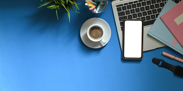 Image vue de dessus du smartphone à écran blanc blanc mettant sur un bureau de travail coloré entouré de stylos marqueurs, cahiers, smartwatch, tasse à café, porte-crayon et plante en pot. Espace de travail encombré.