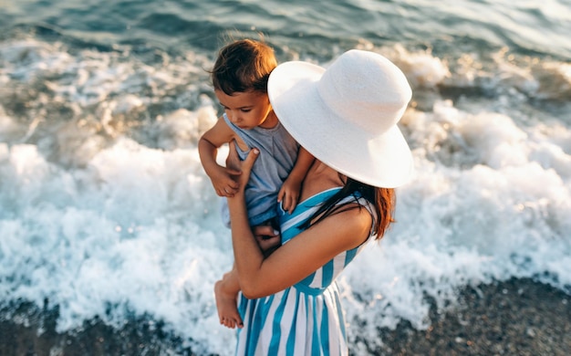 Image vue de dessus horizontale d'une jolie jeune mère jouant avec sa fille au bord de la mer portant une robe à rayures bleues et un fond de chapeau blanc Femme jouant avec une petite fille joyeuse au coucher du soleil sur l'océan