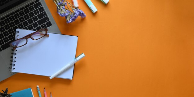 Image vue de dessus d'un ordinateur portable mettant sur un bureau de travail coloré entouré d'un ordinateur portable, de lunettes, d'un stylo, de fleurs, de marqueurs et d'un crayon. Concept d'espace de travail ordonné.