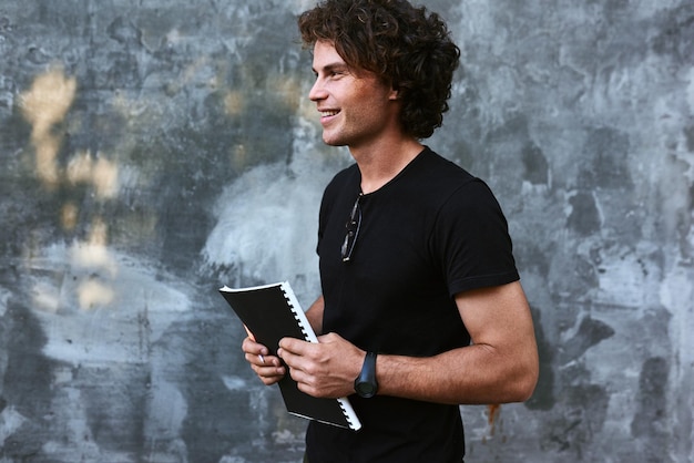 Image vue latérale extérieure d'un homme heureux aux cheveux bouclés écrivant des avis sur papier et se préparant aux examens portant un t-shirt noir posant sur un mur en béton