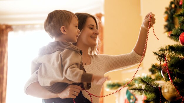 Images 4k d'une mère souriante heureuse avec son enfant mettant des perles et des guirlandes colorées sur l'arbre de Noël. Famille s'amusant et s'amusant pendant les vacances d'hiver et les célébrations.