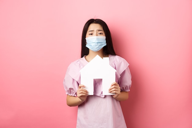 Immobilier et concept de pandémie. Mendicité fille asiatique montrant la découpe de la maison en papier, portant un masque médical, demandant un appartement, debout sur fond rose.