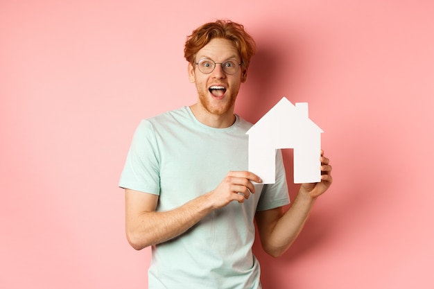 Immobilier. Homme rousse gai, achat d'une propriété, montrant la découpe de maison en papier et souriant étonné, debout sur fond rose.