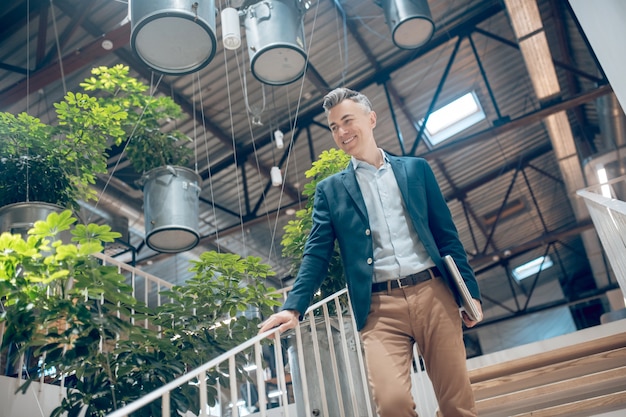 Impression positive. Homme souriant satisfait avec un ordinateur portable descendant les escaliers à l'intérieur avec un intérieur élégant et moderne