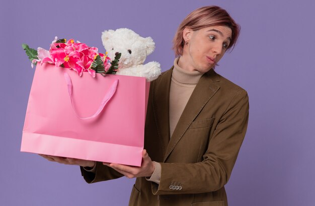 Impressionné jeune bel homme tenant un sac cadeau rose avec des fleurs et un ours en peluche regardant de côté