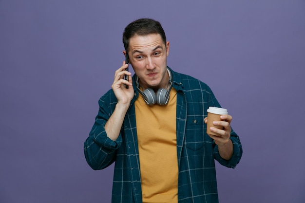 Impressionné jeune étudiant masculin portant des écouteurs autour du cou tenant une tasse de café en papier parlant au téléphone regardant vers le bas isolé sur fond violet