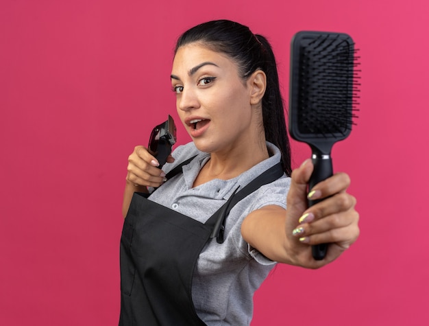 Impressionné jeune femme barbier en uniforme debout dans la vue de profil tenant une tondeuse à cheveux et étirant un peigne isolé sur un mur rose