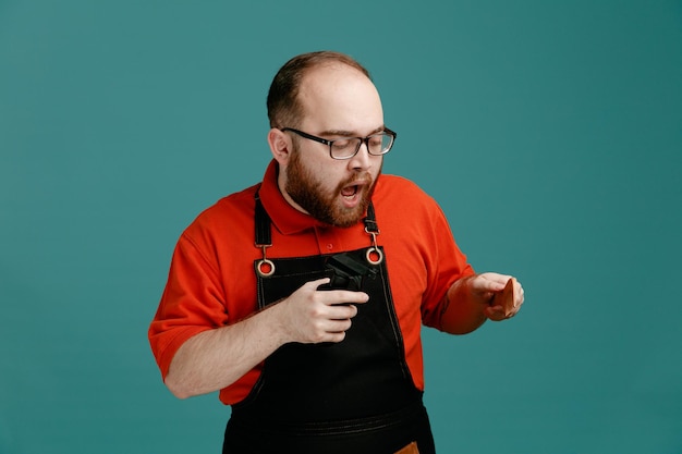 Impressionné jeune homme coiffeur portant des lunettes chemise rouge et tablier de coiffeur tenant un peigne et un fixatif pour cheveux regardant vers le bas isolé sur fond bleu