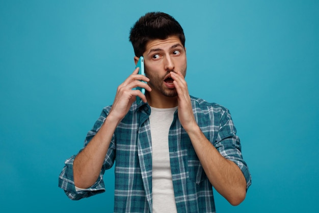 Impressionné jeune homme parlant au téléphone en gardant la main près de la bouche en regardant le côté chuchoter isolé sur fond bleu