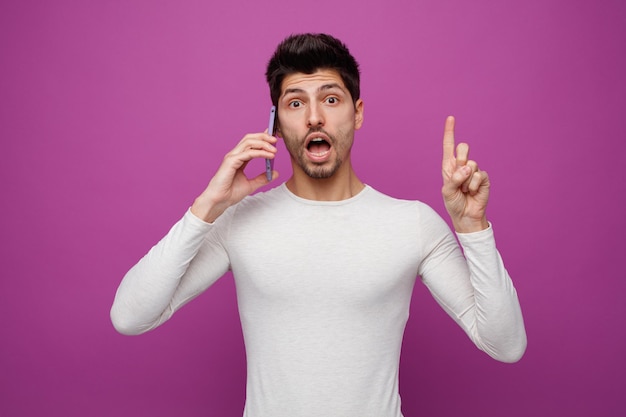 Impressionné jeune homme regardant la caméra pointant vers le haut tout en parlant au téléphone isolé sur fond violet