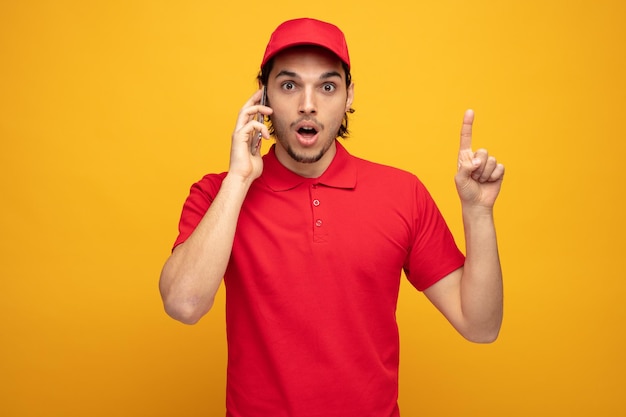 impressionné jeune livreur portant l'uniforme et la casquette regardant la caméra pointant vers le haut tout en parlant au téléphone isolé sur fond jaune