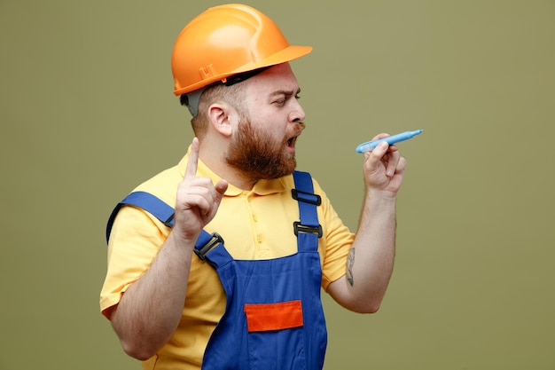 Impressionné tenant marqueur jeune constructeur homme en uniforme isolé sur fond vert