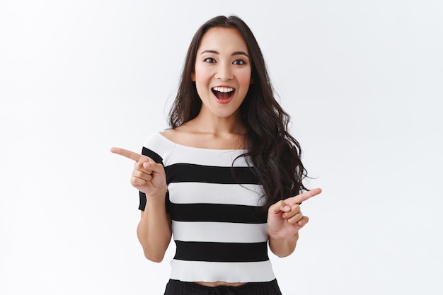 Impressionnée et fascinée, belle fille est-asiatique en t-shirt rayé pointant sur le côté, montrant une publicité à gauche et à droite, souriante amusée et étonnée, regarde la caméra amusée, fond blanc