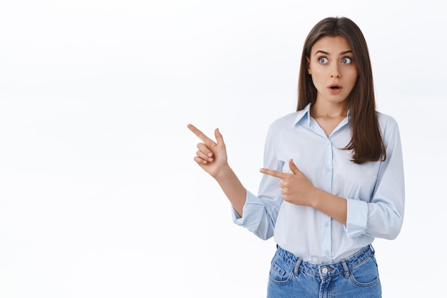 Impressionnée jolie femme sans voix en blouse bleue haletant et regardant fixement quelque chose de cool pointant du doigt la gauche sur la bannière publicitaire du produit montrant de superbes nouvelles fond blanc