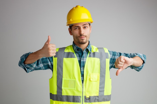 Incertain jeune ingénieur masculin portant un casque de sécurité et un uniforme regardant la caméra montrant les pouces vers le haut et vers le bas isolé sur fond blanc