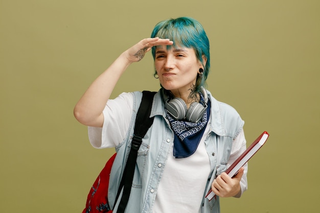 Incertaine jeune étudiante portant des écouteurs et un bandana sur le cou et le sac à dos tenant un carnet de notes en gardant la main devant le front regardant de côté à distance isolé sur fond vert olive