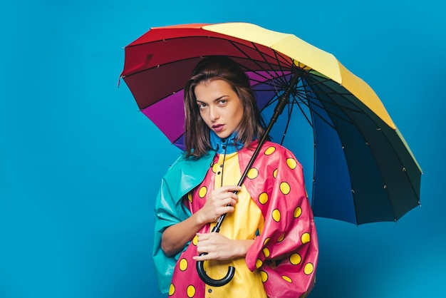 Incroyable belle femme avec un parapluie arc-en-ciel isolé sur fond bleu L'humeur et le temps d'automne sont chauds et ensoleillés et la pluie est possible Les filles en automne se préparent pour la vente d'automne