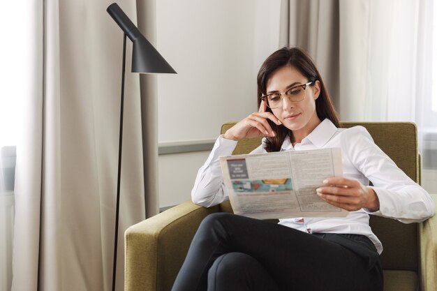 incroyable concentré belle jeune femme d'affaires dans des vêtements de cérémonie à l'intérieur à la maison en lisant le journal.