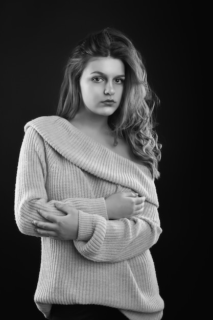 Incroyable femme brune aux longs cheveux bouclés porte un pull tricoté avec des épaules nues posant dans l'ombre. Prise de vue monochrome