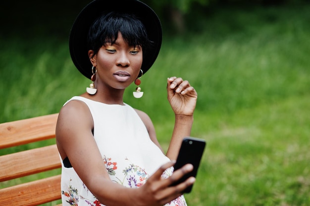 Incroyable femme modèle afro-américaine en pantalon vert et chapeau noir posé sur un banc au parc et faisant selfie sur téléphone mobile
