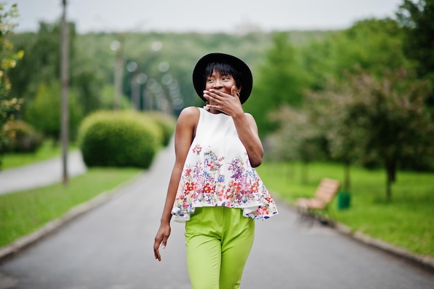 Incroyable femme modèle afro-américaine en pantalon vert et chapeau noir posé avec différentes émotions au parc montre un visage surpris